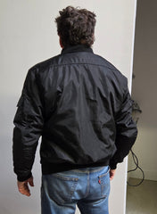Person wearing a black jacket and blue jeans standing against a plain wall.