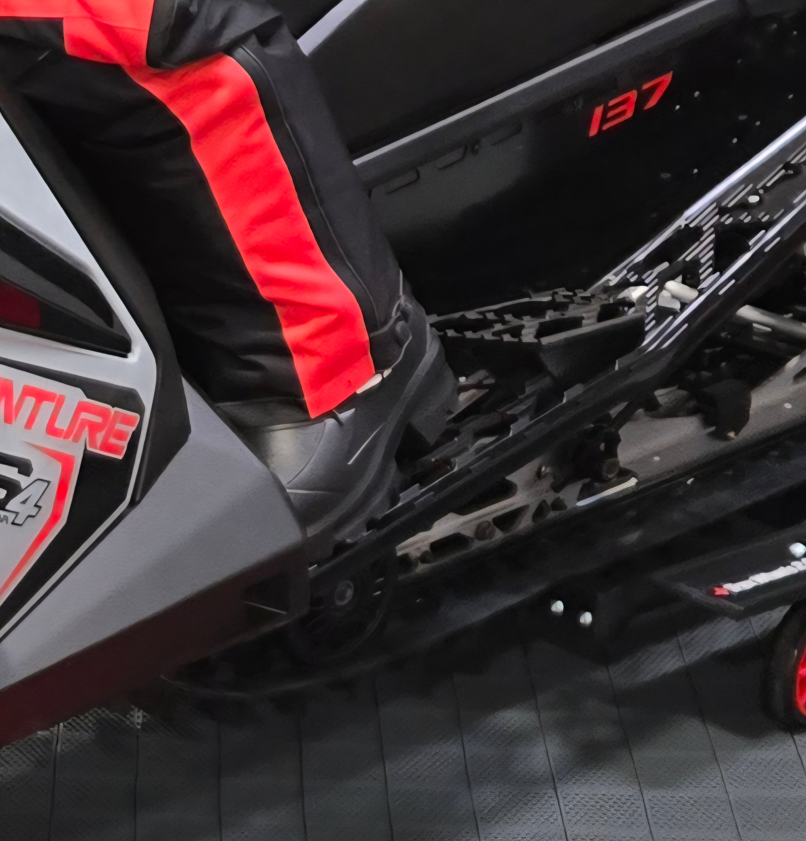 Close-up of a person wearing black and red riding boots on a snowmobile with '137' displayed.
